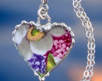 May include: A silver necklace with a heart-shaped pendant. The pendant has a floral design in purple, pink, green, and white, set in a silver frame. The chain is silver with a delicate link design.
