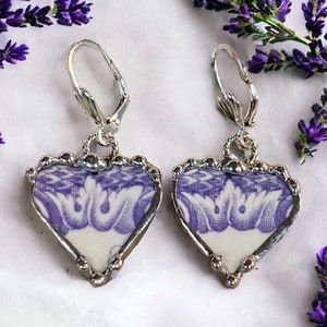 Puede incluir: Un par de pendientes de plata en forma de corazón con un diseño floral morado y blanco. Los pendientes están hechos de porcelana vintage y tienen un cierre de gancho de plata.