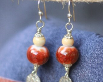 May include: A pair of dangle earrings with silver-tone hooks. Each earring features a cream-colored bead, a large red and orange speckled bead, and a silver-tone, hammered-metal charm. The earrings are suspended from a natural-colored twine.