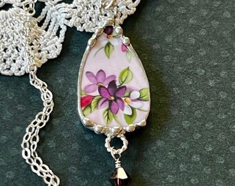 May include: A silver chain necklace with a teardrop-shaped pendant featuring a vintage floral design in shades of pink, purple, and green. A small purple crystal dangles from the bottom of the pendant.