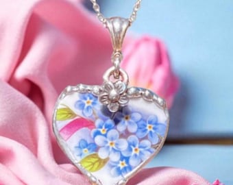 May include: A heart-shaped pendant necklace with a silver chain. The pendant features a floral design of blue forget-me-nots on a white background, framed by a silver border. Pink and blue background.