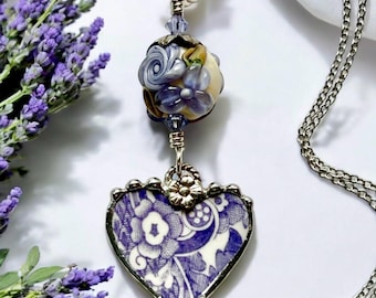 May include: A silver chain necklace with a heart-shaped pendant. The pendant is made of blue and white porcelain with a floral pattern. The necklace also features a round bead with purple and white flowers.