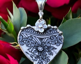 Può includere: Una collana con catena d'argento con un ciondolo a forma di cuore. Il ciondolo è realizzato con un piatto di porcellana vintage con un motivo floreale bianco e nero. Un piccolo ciondolo a forma di fiore d'argento è attaccato alla parte superiore del ciondolo.