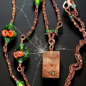 May include: Copper-toned necklace with a Halloween theme. The necklace features a rectangular pendant with a spider and web design, green and orange glass beads, and a chain. The necklace is displayed on a black background.