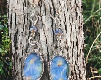 May include: A pair of silver earrings with blue flower accents. The earrings are made with wire and have a delicate, floral design.