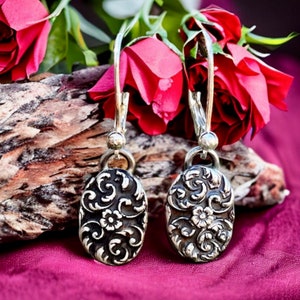 Puede incluir: Un par de pendientes de plata con un diseño floral. Los pendientes son de forma ovalada y tienen un patrón delicado e intrincado. Los pendientes cuelgan de un gancho de plata.