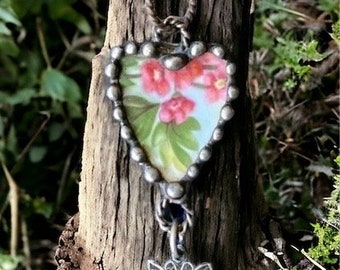 Peut inclure: Un collier pendentif en forme de cœur avec un motif floral. Le pendentif présente un fond bleu clair avec des fleurs roses et des feuilles vertes. Le pendentif est encadré de métal argenté et est suspendu à une chaîne argentée. Un petit papillon est suspendu en dessous.