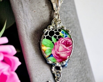 May include: A silver necklace with a round pendant featuring a black and white floral pattern with a pink rose. The pendant is attached to a silver dragonfly charm.