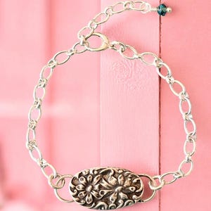 May include: Silver-toned bracelet featuring an oval centerpiece with a floral design. The bracelet has an adjustable chain and a lobster clasp. A small blue bead accents the end of the chain. The bracelet is displayed against a pink background.