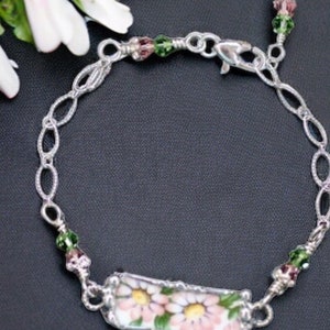 Puede incluir: Un brazalete de cadena de plata con un dije rectangular que presenta un diseño floral rosa y blanco. El dije está rodeado de cuentas verdes y rosas.