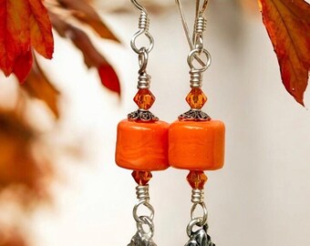 May include: A pair of orange earrings with silver-colored accents. Each earring features a cylindrical orange bead, orange crystal beads, and a silver-colored, diamond-shaped charm. The earrings are suspended from silver-colored hooks.