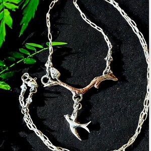 May include: A silver chain necklace with a silver and copper branch pendant with a small bird charm hanging from it. The branch has leaves and the bird is in flight.