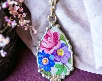 May include: A delicate gold chain necklace with a teardrop-shaped pendant featuring a vintage floral design. The pendant is surrounded by a silver beaded border and has a pink, blue, and purple floral pattern.