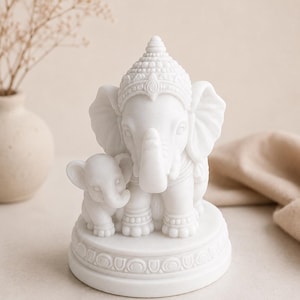 May include: A white matte figurine of a large elephant standing next to a smaller elephant calf. The larger elephant wears an ornate headpiece and jewelry. Both figures are positioned on a circular base featuring a decorative carved border.