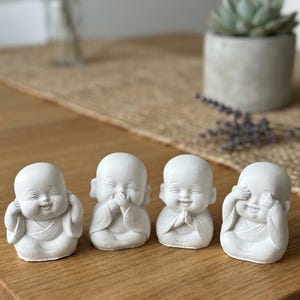 May include: A set of four small, white, matte-finish figurines depicting young monks in seated positions. Each figure displays a different gesture: one covering its ears, one covering its mouth, one with hands in a prayer position, and one covering its eyes.