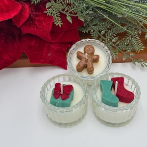 May include: Three small, clear glass votive candle holders with white wax. Each candle has a decorative topping: a gingerbread man, a gift box with a red bow, and a green stocking with a red stocking. Festive holiday decor.