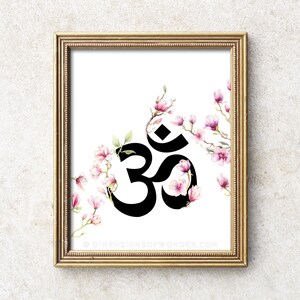 May include: A black and white Om symbol surrounded by pink watercolour flowers on a white background. The image is framed in a gold frame.