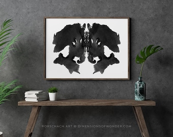 Op de afbeelding: Een ingelijst poster met een zwarte en witte inktvlekprint, hangend aan een grijze muur boven een houten tafel met een potplant en een vaas met een groene plant.