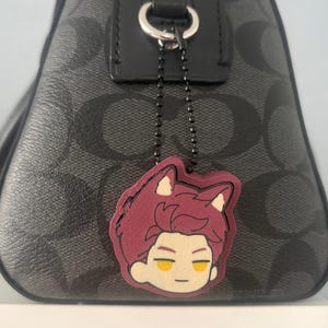 K&ouml;nnte beinhalten: Eine schwarze Handtasche mit grauem Logomuster verf&uuml;gt &uuml;ber einen silbernen Rei&szlig;verschluss und eine schwarze Kugelkette. An der Kette h&auml;ngt ein Gummi-Anh&auml;nger, der einen stilisierten Charakterkopf mit kastanienbraunem Haar und gelben Augen zeigt.