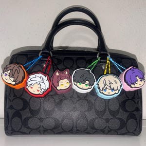 K&ouml;nnte beinhalten: Eine schwarze Handtasche mit wiederkehrendem dunkelgrauem Logomuster und zwei Tragegriffen. Sechs runde Gummi-Anh&auml;nger mit stilisierten Cartoon-Charaktergesichtern in Orange, Rot, Kastanienbraun, Wei&szlig;, Hellblau und Pink h&auml;ngen an den Griffen.