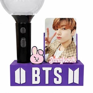 Puede incluir: Soporte de exhibición morado de madera con el tema de BTS, con letras y logotipo blancos. El soporte contiene un palo de luz, una tarjeta fotográfica de una persona y un pequeño personaje de dibujos animados. El soporte tiene un asa y un agujero circular.