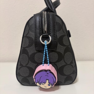 K&ouml;nnte beinhalten: Eine schwarze Handtasche mit grauem Monogramm-Muster und schwarzem Lederbesatz, ausgestattet mit einem silbernen Rei&szlig;verschluss. Eine hellblaue Kugelkette befestigt einen rosa, lila und wei&szlig;en Gummi-Anh&auml;nger mit einem Cartoon-Charakterkopf am Ring.