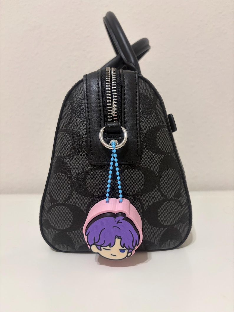 K&ouml;nnte beinhalten: Eine schwarze Handtasche mit grauem Monogramm-Muster und schwarzem Lederbesatz, ausgestattet mit einem silbernen Rei&szlig;verschluss. Eine hellblaue Kugelkette befestigt einen rosa, lila und wei&szlig;en Gummi-Anh&auml;nger mit einem Cartoon-Charakterkopf am Ring.