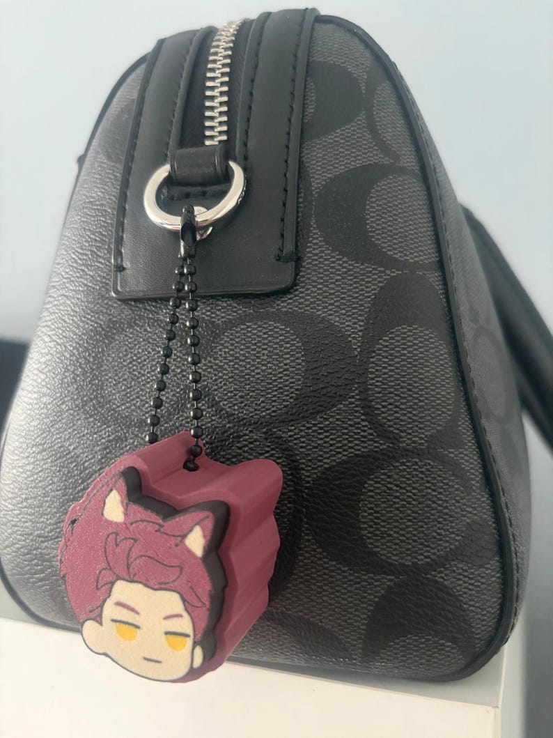 K&ouml;nnte beinhalten: Eine dunkelgraue Handtasche mit einem sich wiederholenden schwarzen Logomuster, einem silbernen Metallrei&szlig;verschluss und einem schwarzen Lederbesatz. Ein kleiner, kastanienbrauner Gummi-Anh&auml;nger in Form eines Anime-Charakterkopfes mit Katzenohren.