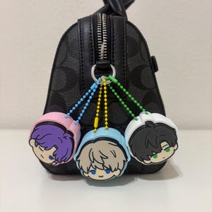 K&ouml;nnte beinhalten: Eine schwarze Handtasche mit Muster, einem silbernen Rei&szlig;verschluss und einem silbernen Metallring. An dem Ring h&auml;ngen drei runde Anh&auml;nger mit Comic-Gesichtern in Rosa, Hellblau und Wei&szlig;, die jeweils an einer farbigen Kugelkette befestigt sind.