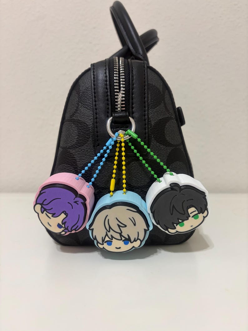 K&ouml;nnte beinhalten: Eine schwarze Handtasche mit Muster, einem silbernen Rei&szlig;verschluss und einem silbernen Metallring. An dem Ring h&auml;ngen drei runde Anh&auml;nger mit Comic-Gesichtern in Rosa, Hellblau und Wei&szlig;, die jeweils an einer farbigen Kugelkette befestigt sind.