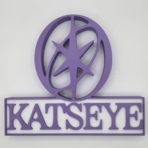 Puede incluir: Un letrero morado con el texto "KATSEYE" en letras blancas. Encima del texto hay un diseño circular morado con forma de estrella en el interior. El letrero es probablemente para un negocio o marca.