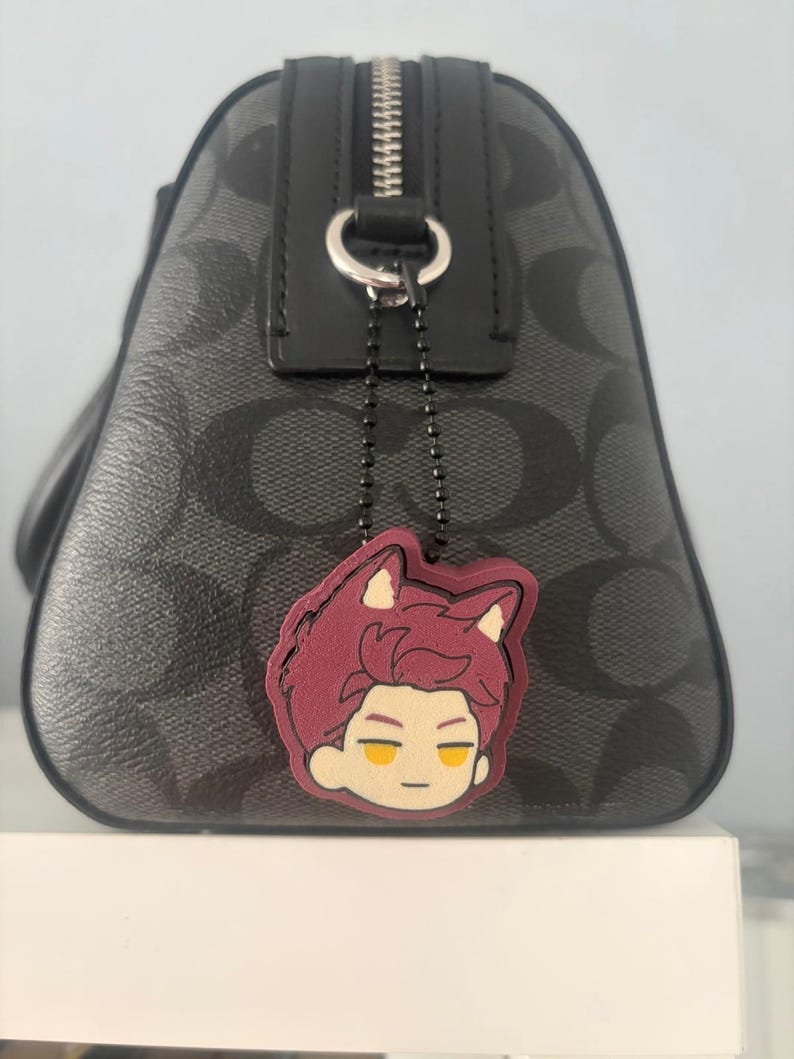 K&ouml;nnte beinhalten: Eine schwarze Handtasche mit grauem Logomuster verf&uuml;gt &uuml;ber einen silbernen Rei&szlig;verschluss und eine schwarze Kugelkette. An der Kette h&auml;ngt ein Gummi-Anh&auml;nger, der einen stilisierten Charakterkopf mit kastanienbraunem Haar und gelben Augen zeigt.