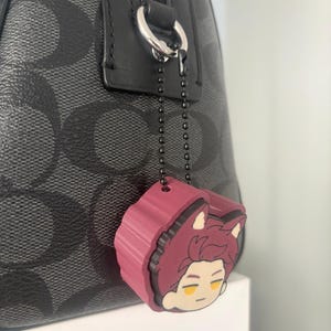 K&ouml;nnte beinhalten: Eine schwarze Handtasche mit grauem Logomuster verf&uuml;gt &uuml;ber einen silbernen Rei&szlig;verschlussanh&auml;nger an einer dunklen Metallkugelkette. Ein kastanienbrauner Gummi-Anh&auml;nger in Form eines Anime-Charakterkopfes mit spitzen Ohren h&auml;ngt daran.