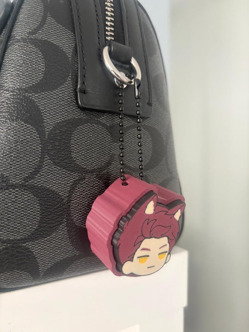 K&ouml;nnte beinhalten: Eine schwarze Handtasche mit grauem Logomuster verf&uuml;gt &uuml;ber einen silbernen Rei&szlig;verschlussanh&auml;nger an einer dunklen Metallkugelkette. Ein kastanienbrauner Gummi-Anh&auml;nger in Form eines Anime-Charakterkopfes mit spitzen Ohren h&auml;ngt daran.