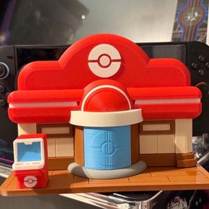 May include: A red and white 3D-printed model of a Pokémon Center, with a blue and white design on the front. A small red and white game console model is in front of the center. A black game console is in the background.