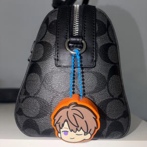 K&ouml;nnte beinhalten: Eine schwarz-grau gemusterte Handtasche mit silbernem Rei&szlig;verschluss und schwarzem Lederbesatz. Ein leuchtend orangefarbener Gummi-Anh&auml;nger, der eine Zeichentrickfigur mit braunen Haaren und einem violetten Auge darstellt, ist befestigt.