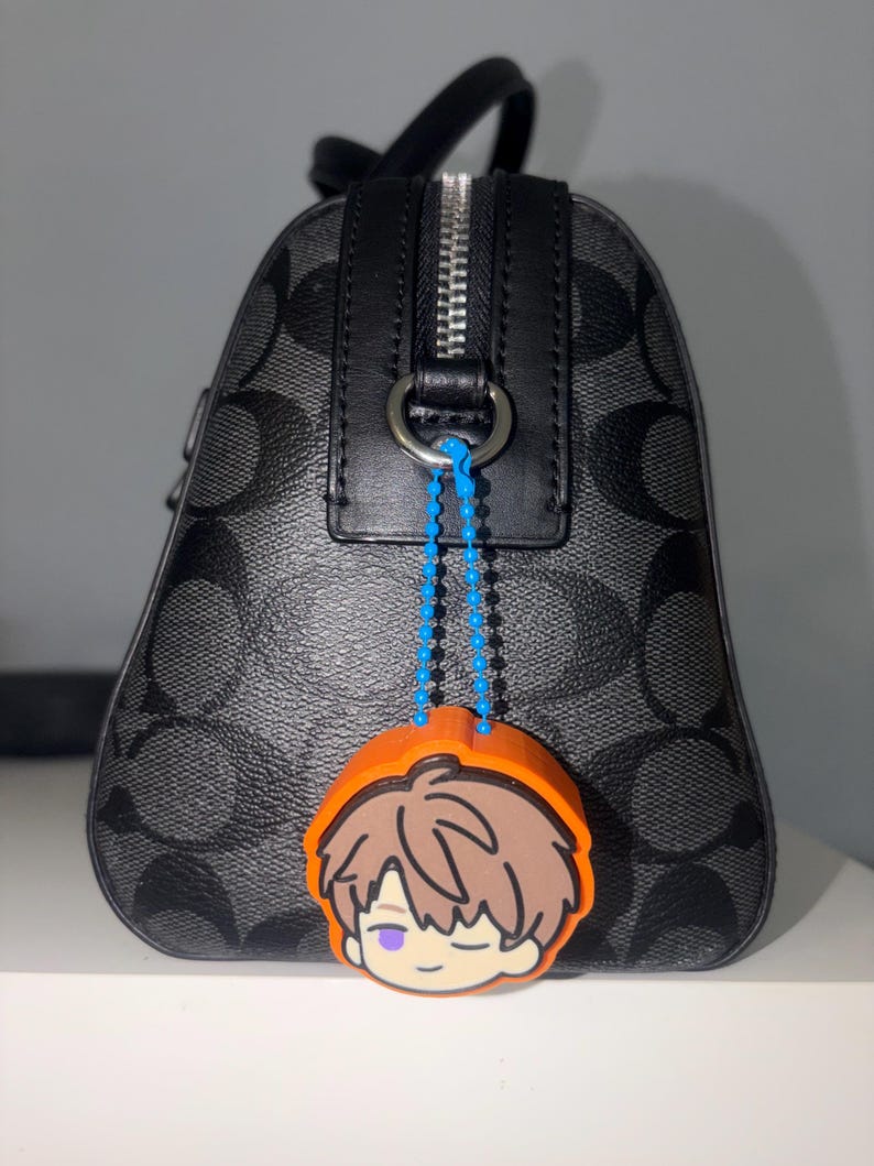 K&ouml;nnte beinhalten: Eine schwarz-grau gemusterte Handtasche mit silbernem Rei&szlig;verschluss und schwarzem Lederbesatz. Ein leuchtend orangefarbener Gummi-Anh&auml;nger, der eine Zeichentrickfigur mit braunen Haaren und einem violetten Auge darstellt, ist befestigt.