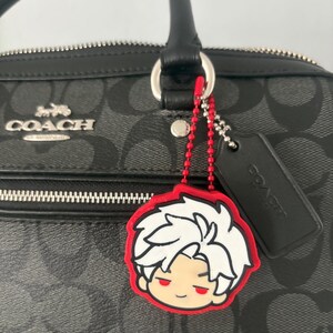 K&ouml;nnte beinhalten: Eine schwarze Coach-Handtasche mit grauem Monogramm-Muster, silberfarbenem Metall-Logo, schwarzem Lederanh&auml;nger und einem rot-wei&szlig;en Gummi-Anh&auml;nger in Form einer Anime-Figur mit stacheligem wei&szlig;em Haar, befestigt an einer roten Kugelkette.