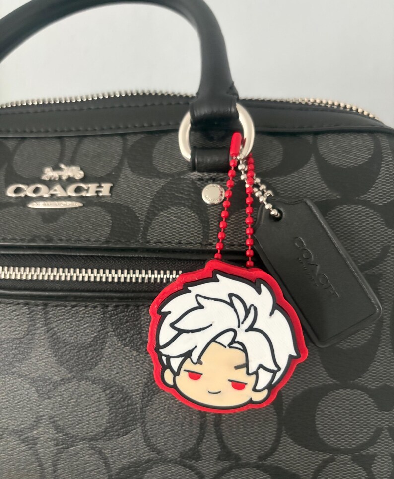 K&ouml;nnte beinhalten: Eine schwarze Coach-Handtasche mit grauem Monogramm-Muster, silberfarbenem Metall-Logo, schwarzem Lederanh&auml;nger und einem rot-wei&szlig;en Gummi-Anh&auml;nger in Form einer Anime-Figur mit stacheligem wei&szlig;em Haar, befestigt an einer roten Kugelkette.