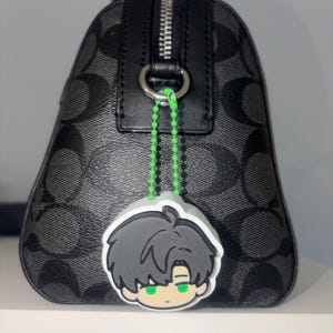 K&ouml;nnte beinhalten: Eine schwarze Handtasche mit grauem Logomuster und silbernem Rei&szlig;verschluss. Eine neongr&uuml;ne Perlenkette befestigt einen Gummi-Anh&auml;nger am Metallring der Tasche. Der Anh&auml;nger zeigt eine Comicfigur mit dunklem Haar und leuchtend gr&uuml;nen Augen.