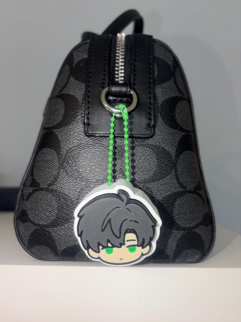 K&ouml;nnte beinhalten: Eine schwarze Handtasche mit grauem Logomuster und silbernem Rei&szlig;verschluss. Eine neongr&uuml;ne Perlenkette befestigt einen Gummi-Anh&auml;nger am Metallring der Tasche. Der Anh&auml;nger zeigt eine Comicfigur mit dunklem Haar und leuchtend gr&uuml;nen Augen.