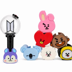May include: A black and clear spherical light stick with a BTS logo inside, accompanied by a collection of seven colourful silicone character covers featuring various faces including a pink rabbit, red heart, blue koala, and yellow-rimmed character.