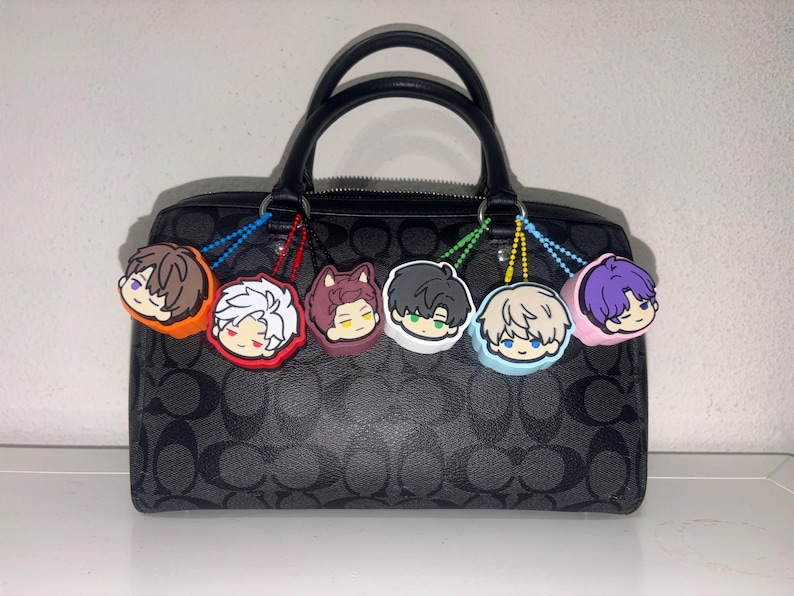 K&ouml;nnte beinhalten: Eine schwarze Handtasche mit wiederkehrendem dunkelgrauem Logomuster und zwei Tragegriffen. Sechs runde Gummi-Anh&auml;nger mit stilisierten Cartoon-Charaktergesichtern in Orange, Rot, Kastanienbraun, Wei&szlig;, Hellblau und Pink h&auml;ngen an den Griffen.