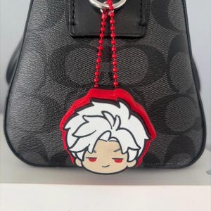 K&ouml;nnte beinhalten: Eine schwarze Handtasche mit grauem Monogramm-Muster verf&uuml;gt &uuml;ber eine rote Kugelkette, die an einem silbernen Ring befestigt ist. An der Kette h&auml;ngt ein Gummi-Anh&auml;nger, der eine Comicfigur mit wei&szlig;en Haaren und roten Augen darstellt.