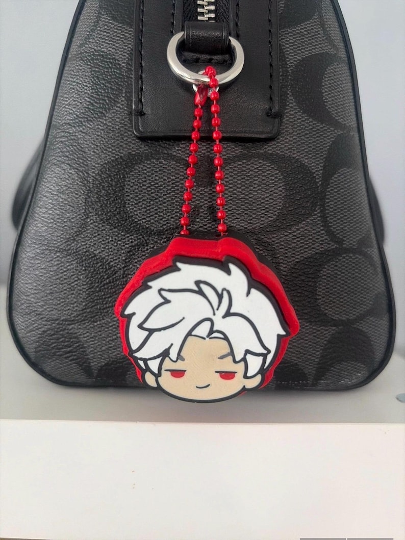 K&ouml;nnte beinhalten: Eine schwarze Handtasche mit grauem Monogramm-Muster verf&uuml;gt &uuml;ber eine rote Kugelkette, die an einem silbernen Ring befestigt ist. An der Kette h&auml;ngt ein Gummi-Anh&auml;nger, der eine Comicfigur mit wei&szlig;en Haaren und roten Augen darstellt.