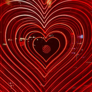 May include: A vibrant, red neon heart design. The image features concentric heart shapes, creating a tunnel effect. The bright red lines contrast against a dark background, with a small, circular detail at the center.