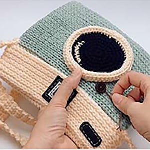 May include: A handmade, knitted bag in shades of beige and teal. The bag features a large, oval-shaped black and cream-colored design on the front, with a button closure. The bag has a strap and a rectangular patch with black stitching.