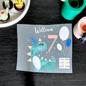 Puede incluir: Tarjeta de cumpleaños gris claro con el nombre "William" y el número "7". La tarjeta presenta un dinosaurio de dibujos animados con un gorro de fiesta, globos y un regalo envuelto. Hay confeti esparcido.