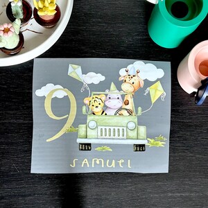 Könnte beinhalten: Ein grauer Aufbügler mit einem hellgrünen Jeep mit einem Tiger, einem Flusspferd und einer Giraffe. Die Zahl 9 und der Name "SAMUEL" sind gelb. Zwei Drachen und Wolken sind ebenfalls vorhanden.