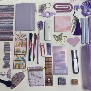 Puede incluir: Una colección de artículos de papelería y accesorios morados, que incluyen un cuaderno, bolígrafos, pegatinas, un estuche y una goma para el pelo. Los artículos están dispuestos sobre una superficie blanca, creando una presentación visualmente atractiva. También hay elementos decorativos.